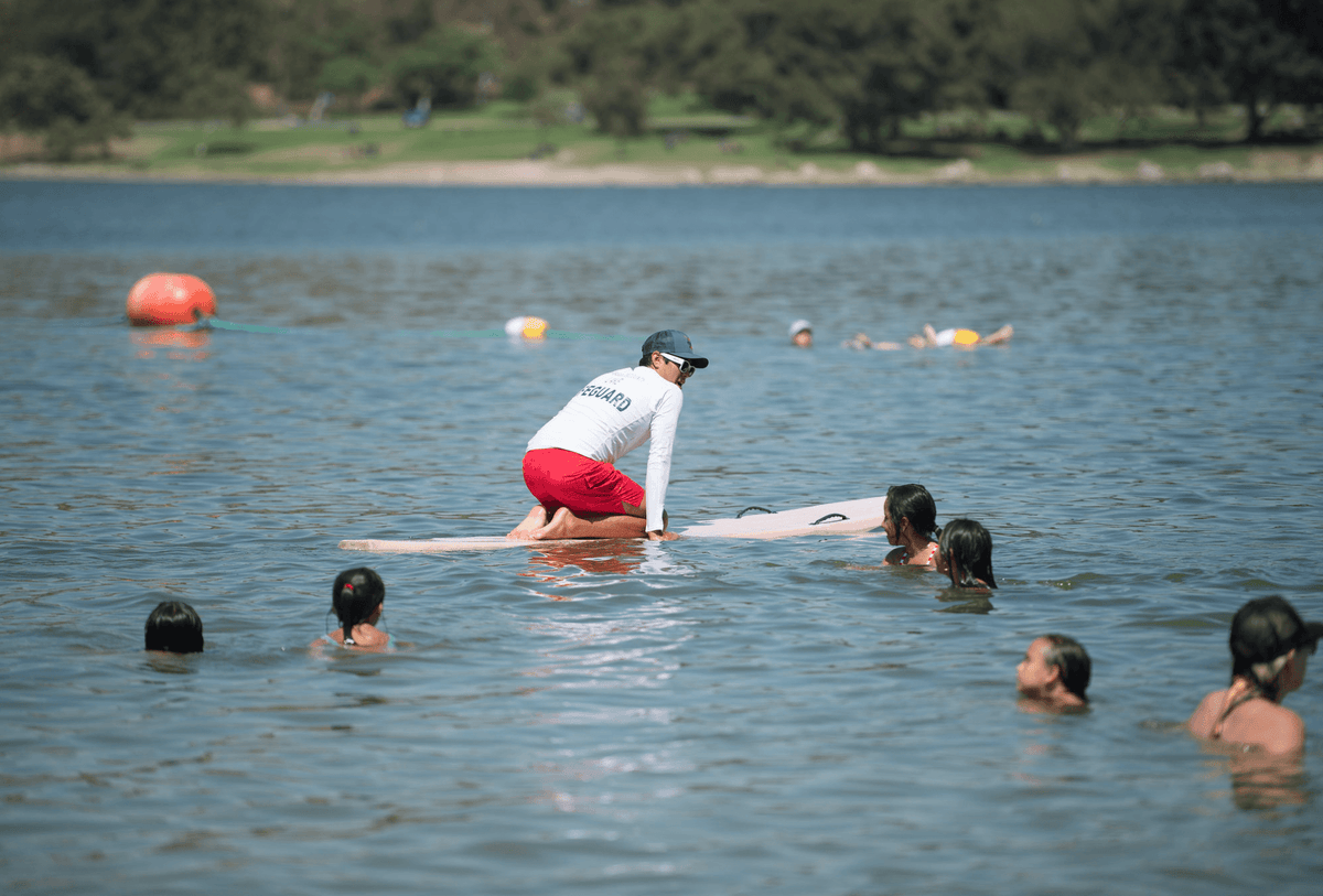 Instructor in red shorts and lifeguard shirt teaching waterfront lifeguard certification course on a lake with students in water. Ideal for lake safety training.